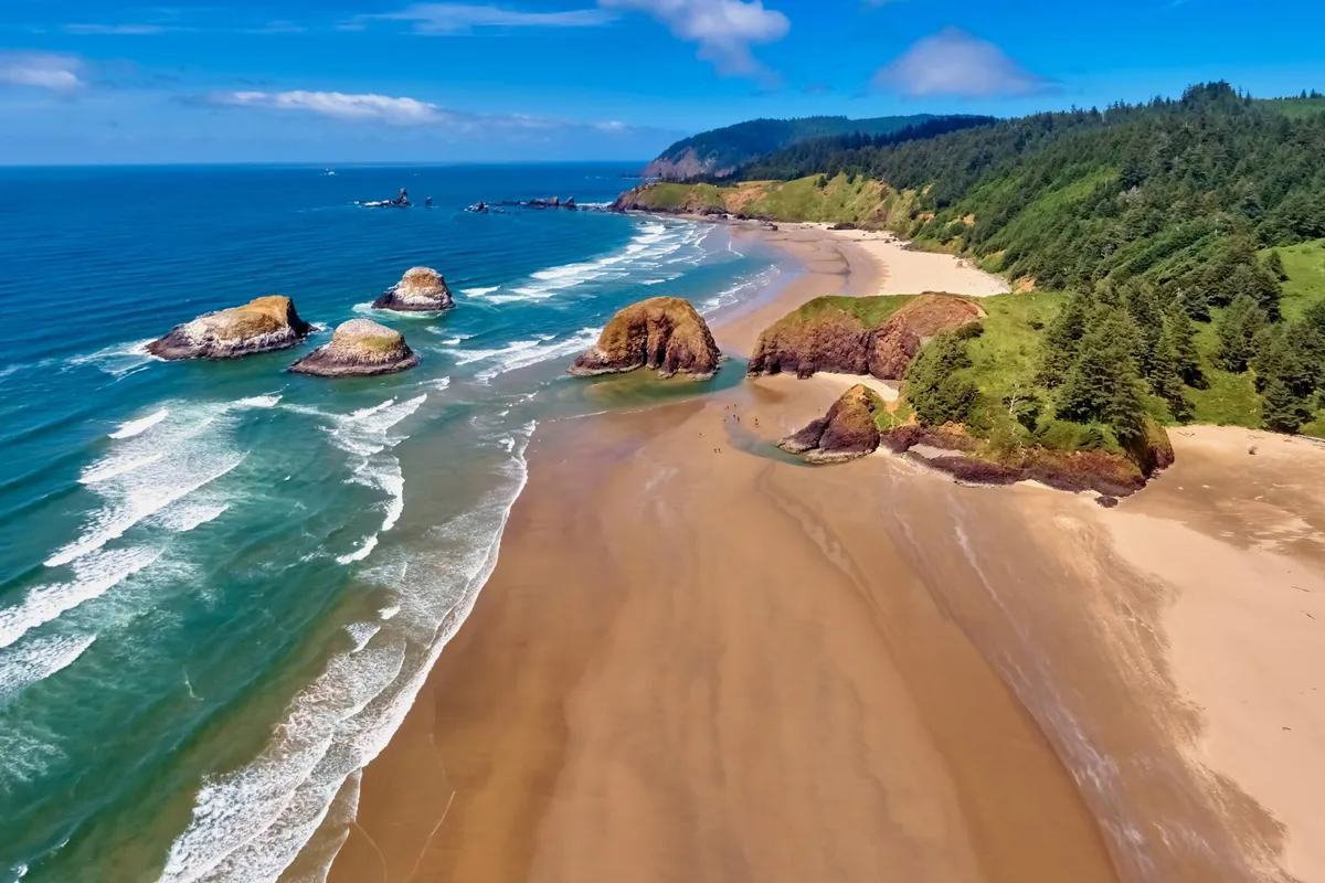 A tranquil exterior scene showing the sweeping sandy beach edged by rocky outcrops and forested hills. Ocean waves lap gently at the shore, inviting exploration and leisurely strolls. The natural setting suggests opportunities for beachcombing, sunsets, and connecting with nature right outside your door.