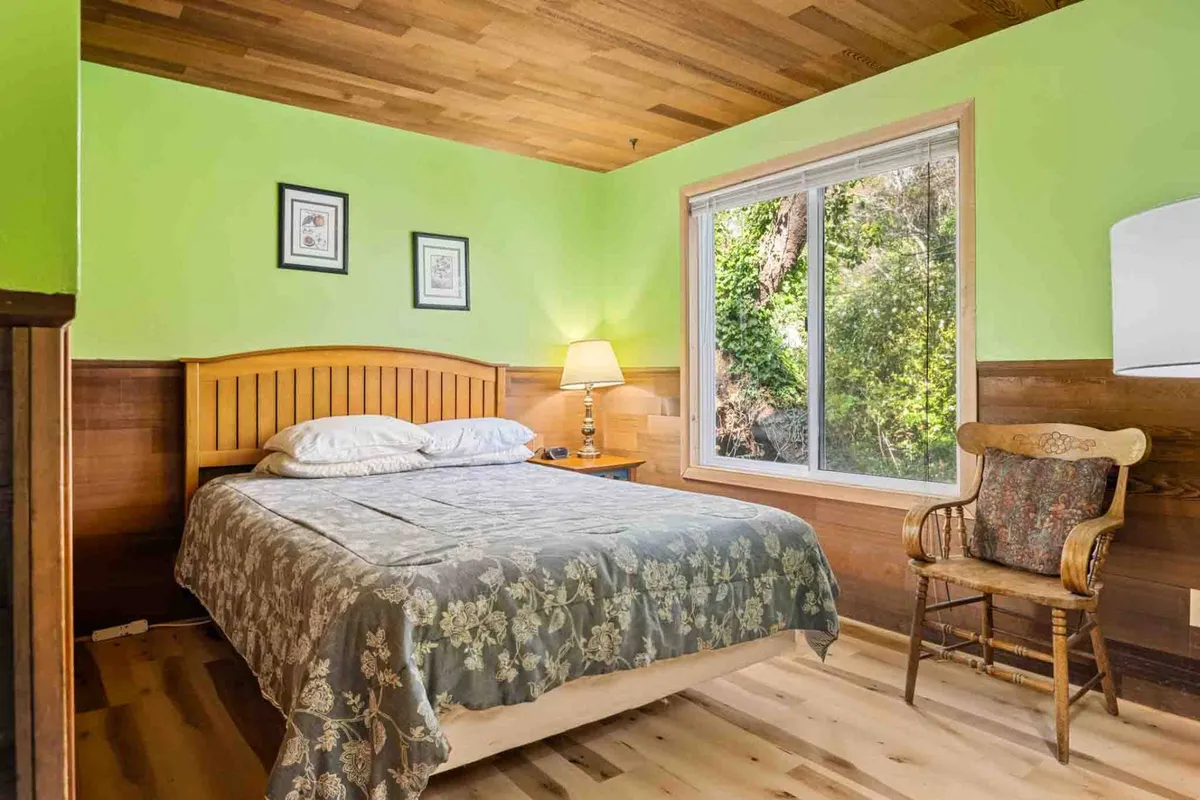 A bright loft-like bedroom featuring a bed, reading chair, lamp, and a window with views outside. The intimate setting and elevated feel suggest a treetop retreat, perfect for privacy and tranquility.
