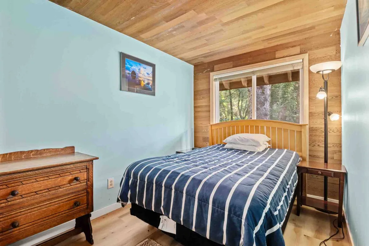 A serene bedroom with a bed featuring striped bedding, a wooden dresser, a lamp next to the bed, framed artwork on the wall, and a window with a view of nature. The setting is calm and uncluttered, ideal for rest.