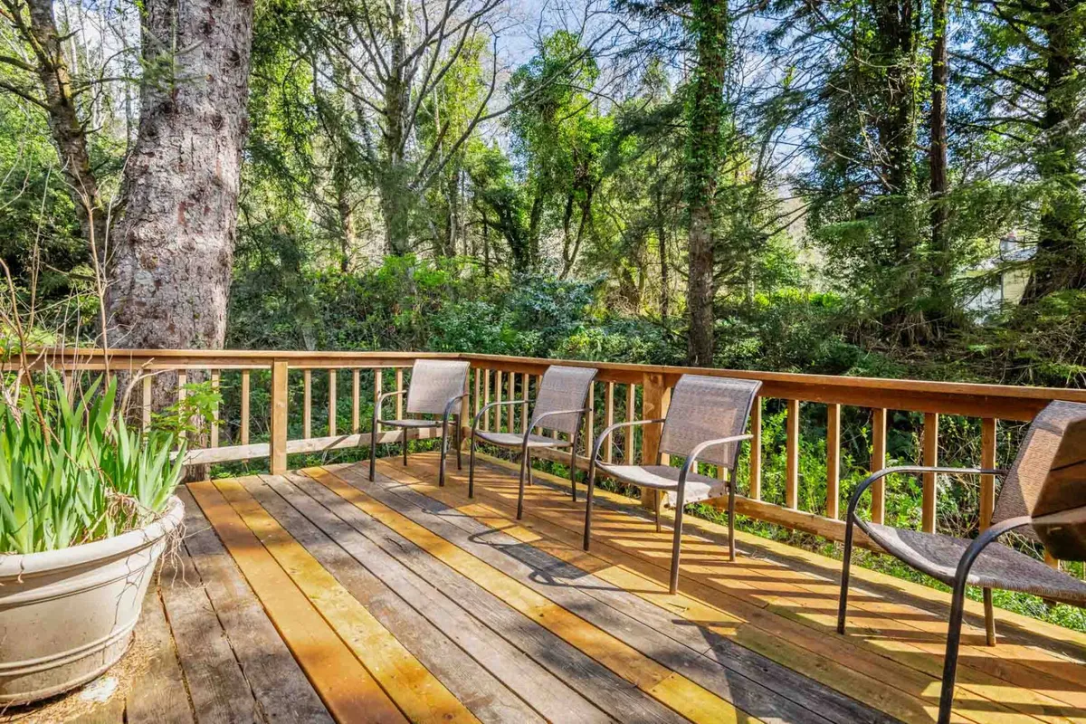 An outdoor wooden deck surrounded by greenery and trees, furnished with comfortable chairs and a planter. The space is inviting for grilling, dining al fresco, or soaking in sunset views, linking the interior experience to the outdoors.