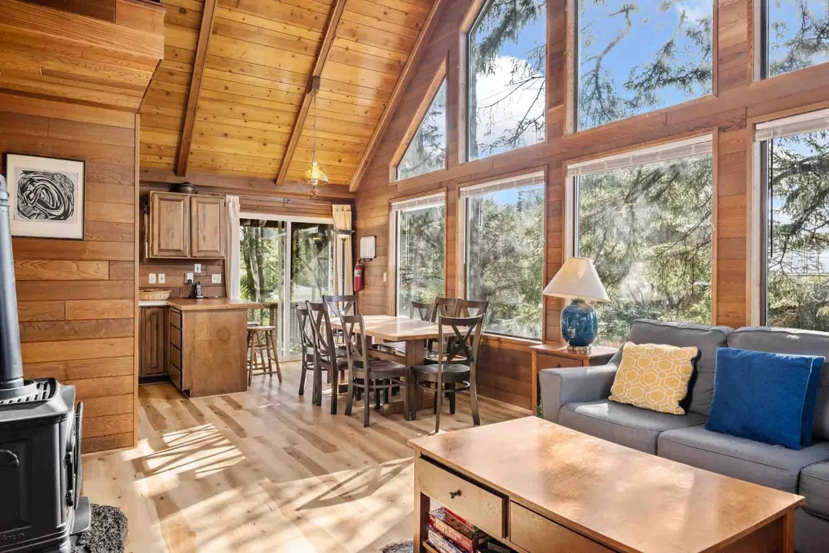 A sunlit open-concept living and dining area features soaring wooden beams, expansive windows that frame Tolovana Park sunsets, a comfy sofa, and a spacious dining table perfect for family meals or game nights.