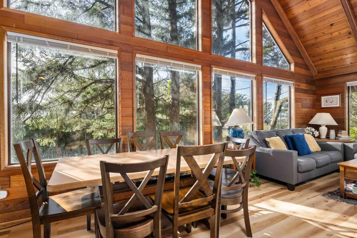 A spacious, open-plan dining area with a large wooden table, big windows letting in natural light, and visible wooden beams. The adjacent living space hints at lively family gatherings and meals enjoyed together, reinforcing the sense of community.