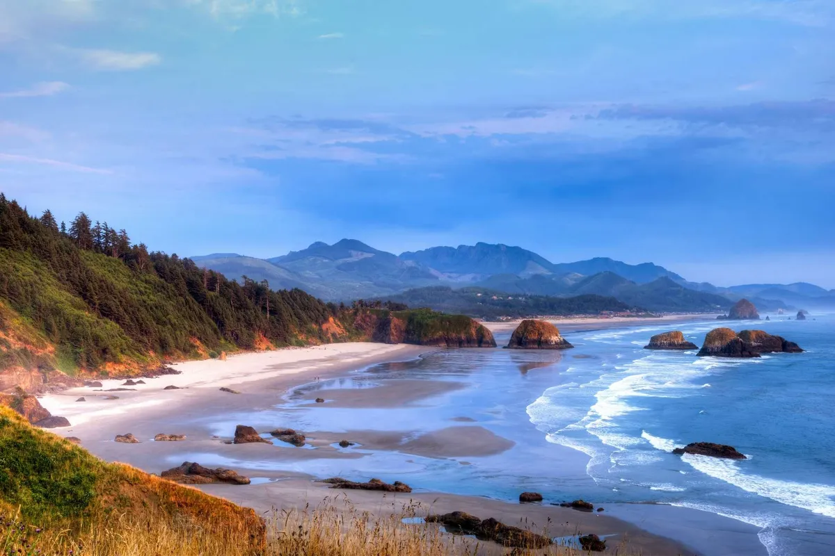 A breathtaking view of Tolovana Beach, featuring golden sands stretching along the shoreline, dramatic rock formations including Haystack Rock in the distance, and gentle waves meeting the shore. In the background, low mountains create a picturesque backdrop. The scene captures people walking along the beach, exploring tide pools, and enjoying the coastal landscape.