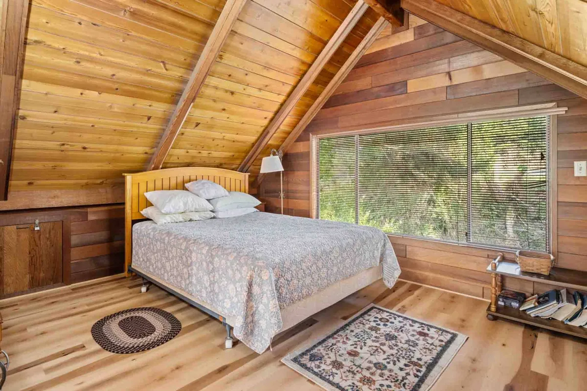 A tranquil bedroom with a wooden bed, textured walls, a cozy rug, and a large window letting in natural light and views of greenery. The setting exudes relaxation and a connection to the outdoors.